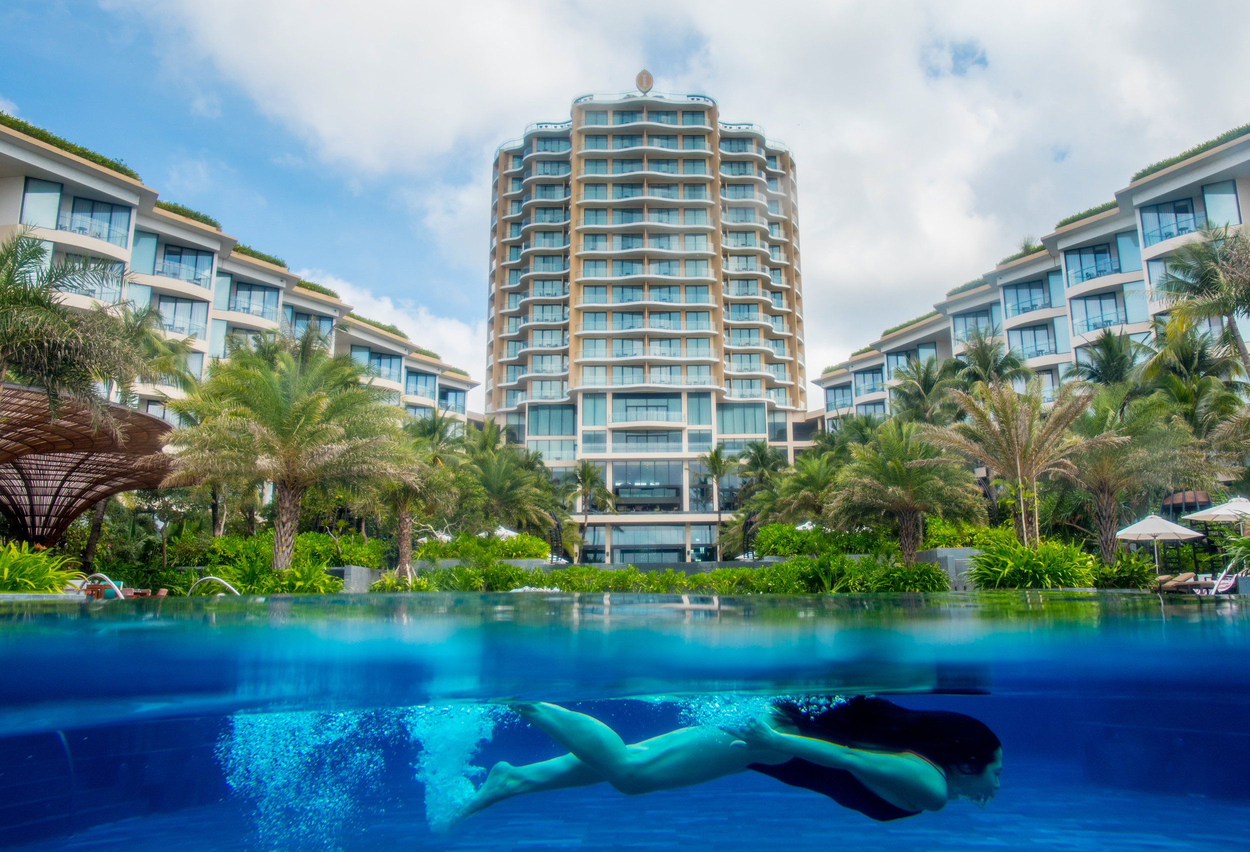 A swimmer glides underwater in a resort pool framed by palms and a tall hotel.