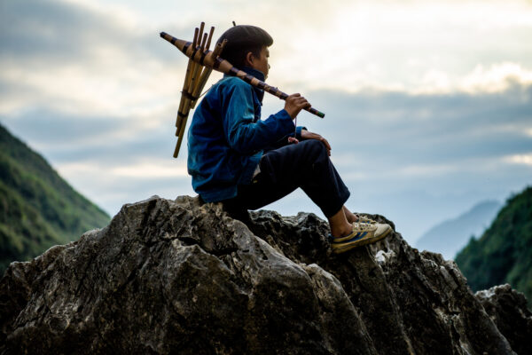 Person sitting on a jagged rock outcrop with a bundle of bamboo wind instruments on their back, overlooking misty mountains at sunset/sunrise.