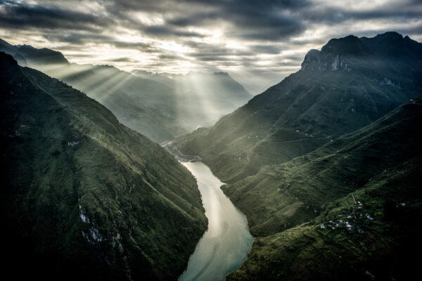 Sunbeams break through the clouds onto a deep green valley with a winding river between rugged mountains.