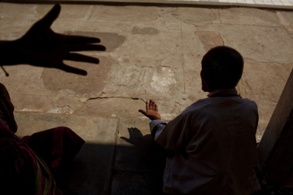Silhouetted child facing a concrete wall, reaching toward a large shadow of a hand on the wall beside them in a sunlit outdoor scene.