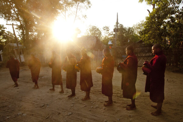 Line of Buddhist monks in saffron robes standing barefoot in a line, each holding a metal alms bowl at sunrise in a dusty village yard.