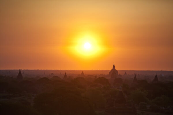 Sunset over a landscape of silhouetted temples and trees, with a bright orange sky and glowing sun centered on the horizon.