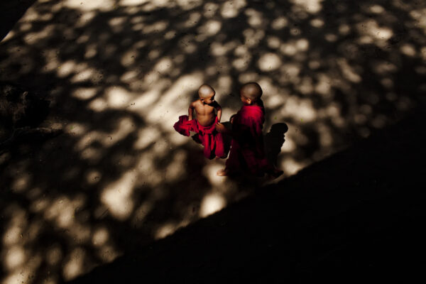 Two young Buddhist monks in red robes walk barefoot on a sunlit path, with dappled shadows around them.