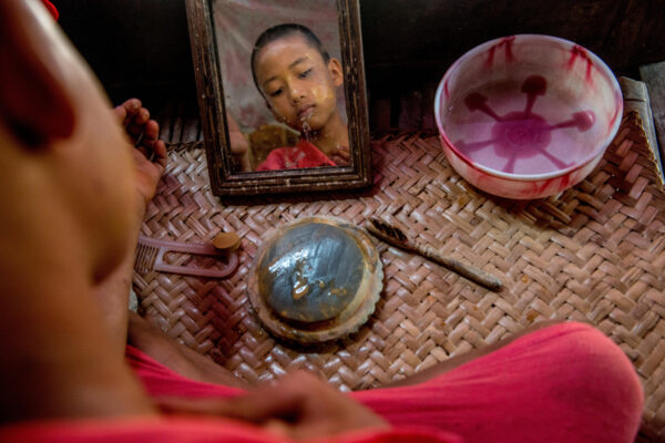 Child sits on a woven mat, looking into a small wooden mirror while a pink basin sits nearby.