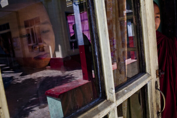 Reflection of a person in a red robe seen through a glass window, with colorful interior and outdoor reflections visible in the panes.