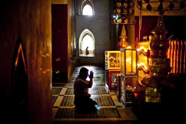 Silhouetted person kneels in prayer inside a dim temple, warm candlelight reflecting off gold ritual objects to the right.