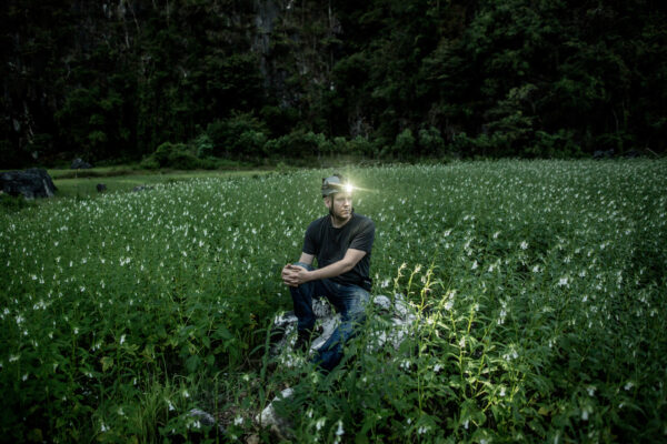 Man sitting on a rock in a field of tall white flowers, wearing a dark shirt and a headlamp that is lit.
