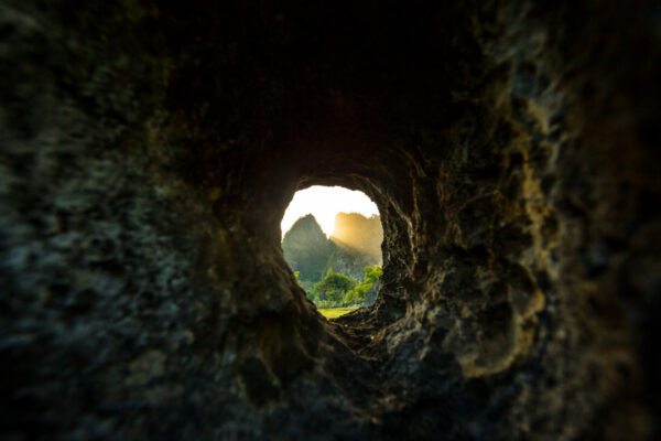 View through a rough rock tunnel opening to a sunlit valley with greenery and distant mountains/karst formations.