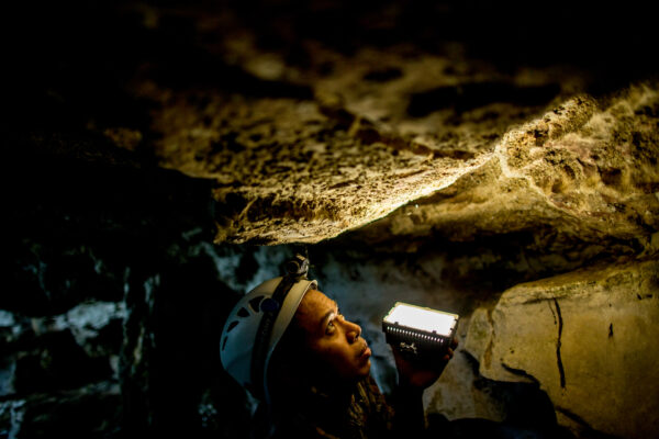 Cave explorer wearing a helmet with a headlamp, holding a bright portable light inside a dark cave.