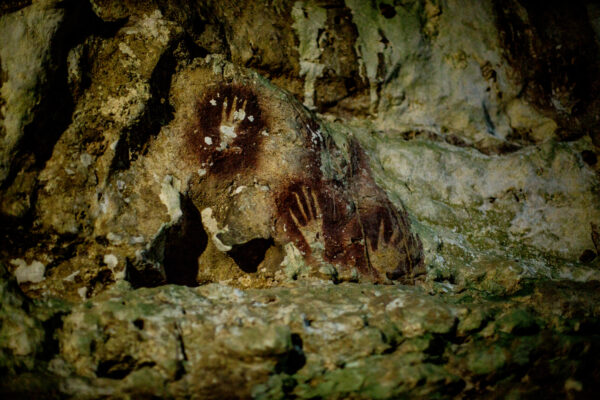 Cave wall with a reddish-brown handprint stencil on rough rock surface.