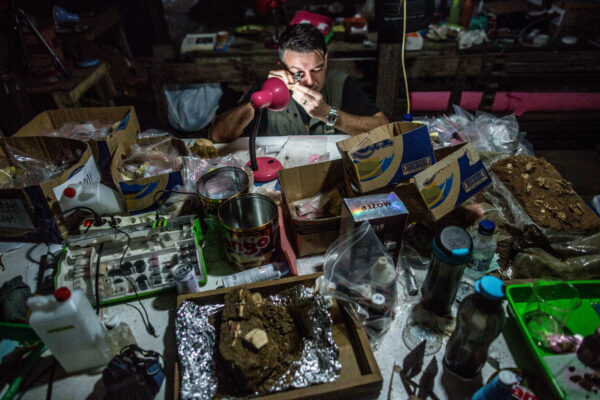 Man in a dim, cluttered workshop inspects a pink tool at a crowded workbench covered with boxes, jars, and soil samples.