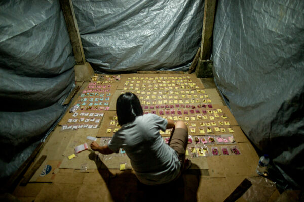 Person sitting on the floor inside a tarp-covered shelter, sorting many small packets laid out in neat rows on cardboard sheets.