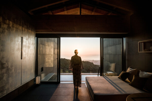 Person in a robe stands at open glass doors, watching a sunset over hills and the sea from a modern bedroom.