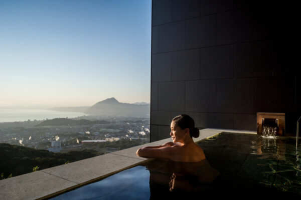 Woman relaxing in an outdoor pool along a modern building, overlooking a coastal city and distant mountains at sunset.”] ,