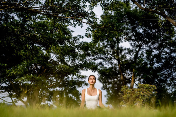 A woman in a white tank top sits cross‑legged in a grassy field, meditating with eyes closed under a canopy of trees.