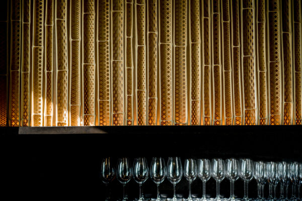 Row of empty wine glasses lined up on a wooden bar shelf with a gold geometric metal backdrop behind them.
