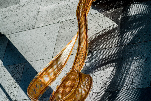 Orange woven ribbon sculpture laid on a speckled gray tiled floor, casting dramatic shadows.