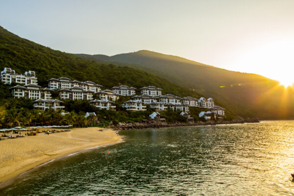 Beach resort with white buildings on a green hillside at sunset, with a calm bay and a small boat on the water.