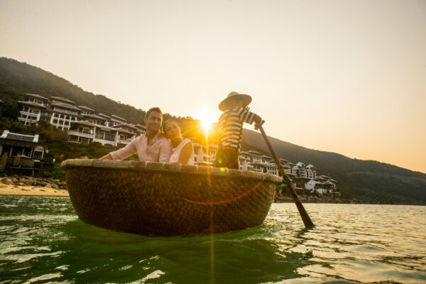 Couple in a woven boat at sunset, with a boatman steering and resort buildings along a hillside behind them.
