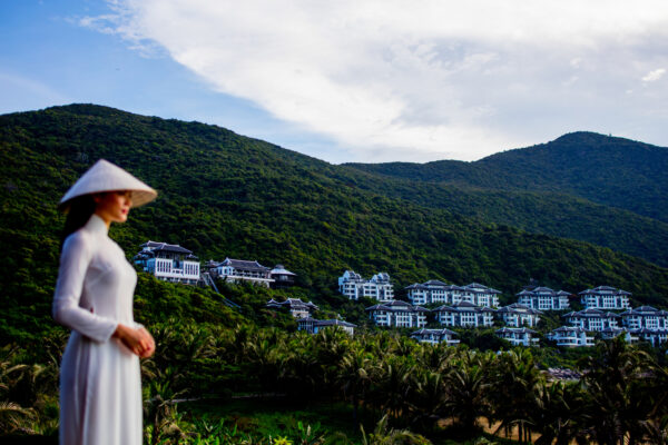Woman in a white áo dài with a conical hat stands in the foreground as a hillside resort village and green mountains loom behind.