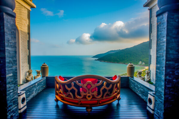 Ornate red-cushioned sofa on a dark wooden deck facing a turquoise bay and green hills under a blue sky, framed by stone walls.