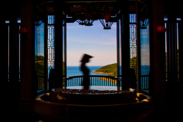 Silhouetted passerby with a hat walking past an open seaside balcony, seen through dark decorative windows with a circular fountain in the foreground.
