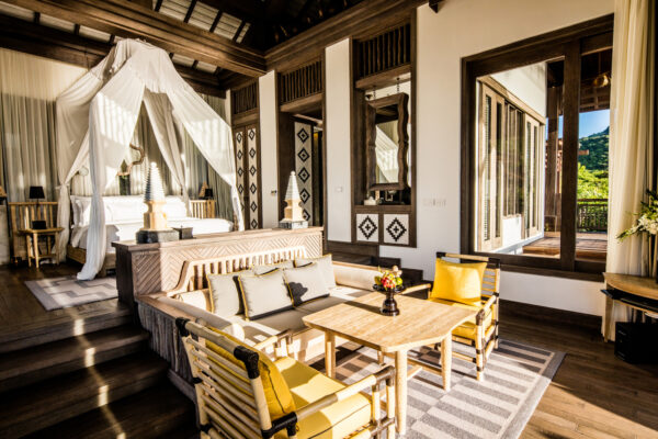 Luxurious bedroom with a four-poster canopy bed draped in white, rich wooden beams, and a seating area with yellow cushions by a sunlit window.Outside view of greenery on a balcony.