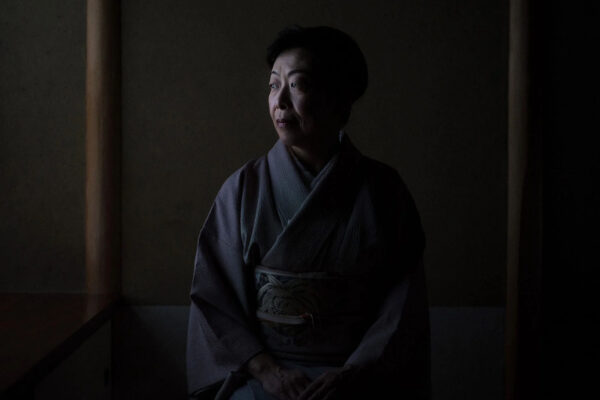 Woman in a traditional kimono sits in a dimly lit room, gazing thoughtfully to the side.
