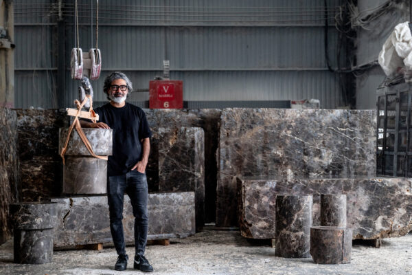 Man with gray hair and beard in a marble workshop, standing beside large, polished marble blocks and a hoist.