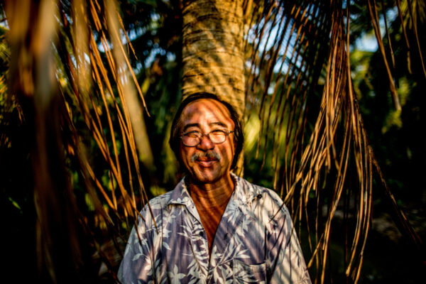 Smiling man with glasses stands among palm fronds, sunlight casting striped shadows on his face and shirt.
