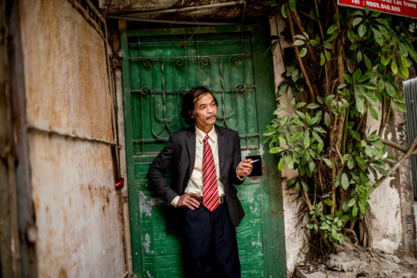 Man in a suit leaning against a weathered green door in an urban alley, smoking a cigarette at the edge of the frame.