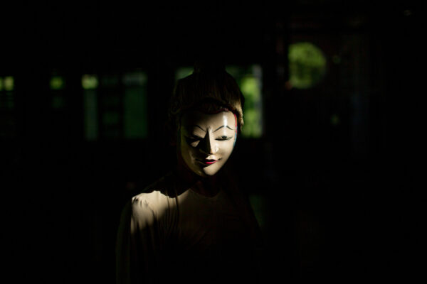 Person wearing a white theatrical mask lit from the side in a dark room; only the face is illuminated with a subtle smile.