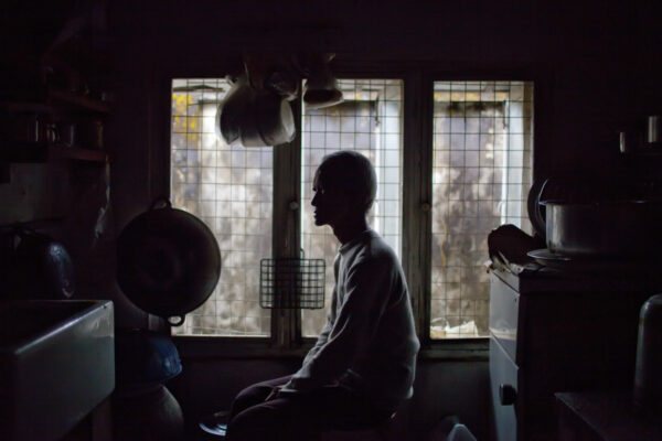 Silhouetted person sitting on a kitchen counter by a barred window, dimly lit with hanging cookware and pots nearby.