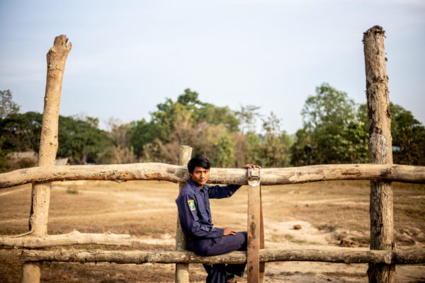 Boy in a blue uniform sits on a weathered wooden fence in a dry, open field with trees in the distance.