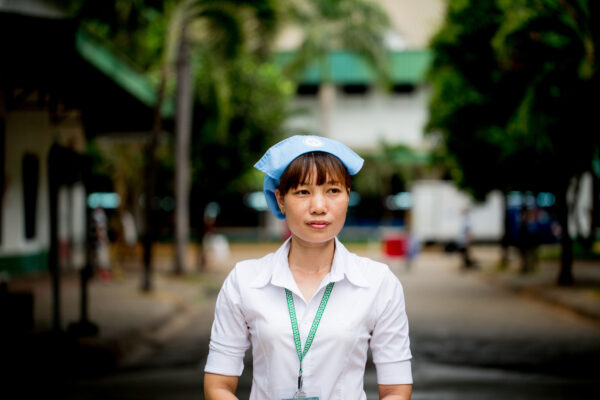 Woman in a white uniform with a light blue cap and lanyard standing on a shaded outdoor path, looking ahead.