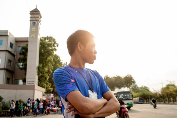 Profile view of a man in a blue shirt with a name badge standing with arms crossed at an outdoor event, with a crowd and buildings in the background.
