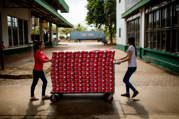 Two women push a wheeled cart stacked with many red-and-white shoe boxes along a paved outdoor path.