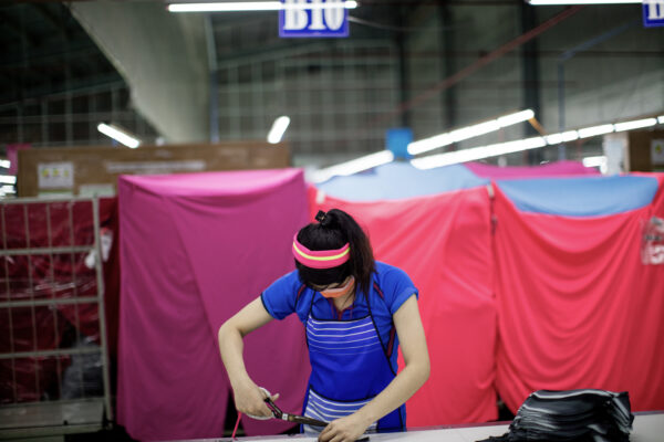 Woman in a blue uniform and pink headband cutting fabric with scissors at a workstation in a textile workshop with pink partitions behind her