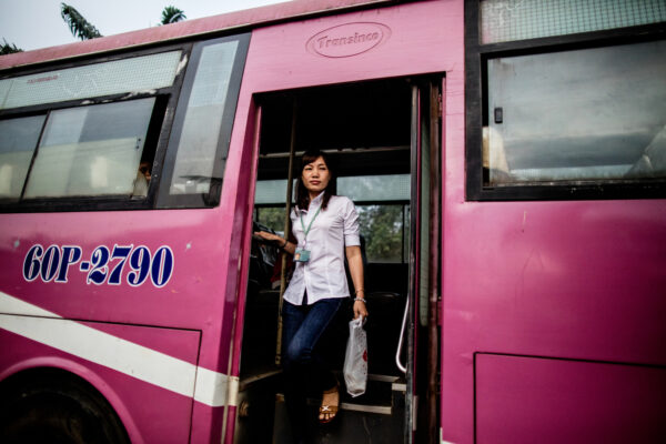 Woman with a lanyard stepping off a pink bus labeled 60P-2790, carrying a plastic bag.