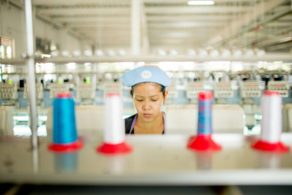 Worker in a light blue cap looking down at a row of colorful thread cones on a textile production line, blurred foreground and background.
