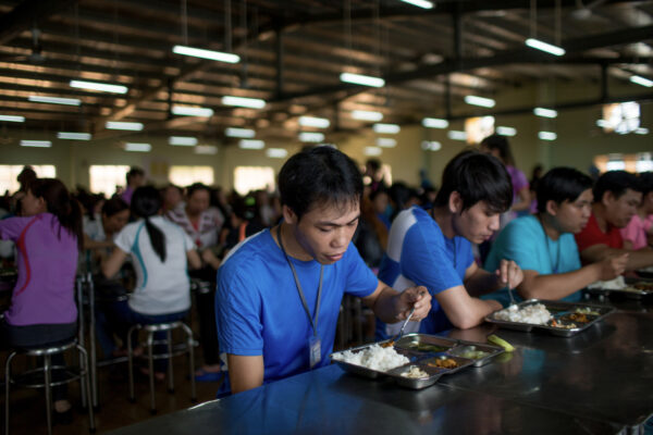 Students in blue shirts eat from metal meal trays in a crowded cafeteria; foreground shows rice and curry dishes on trays while others dine in the background.