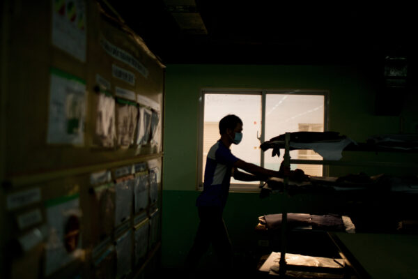 Person wearing a surgical mask walks through a dim room, reaching for items on a shelf near a window.