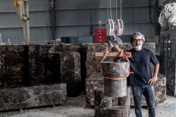 Man with gray beard and glasses stands in a marble workshop beside a suspended cylindrical marble piece strapped with orange lifting slings.