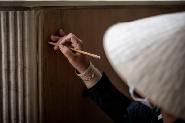 Close-up of a hand holding a pencil and writing on a wooden panel beneath a table, beige sleeve visible