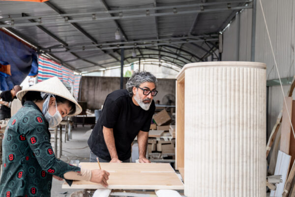Two Woodworkers in a workshop: a woman wearing a conical hat and mask marks wood while a man with gray hair and beard leans over a workbench to inspect a wooden panel.