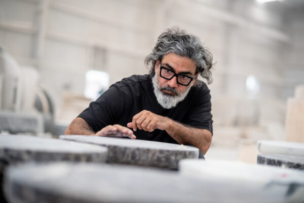 Man with gray hair and beard in a black shirt and glasses leans over a large circular stone slab, inspecting it in a workshop.