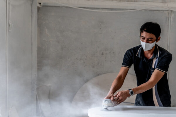 Man wearing a face mask polishes a white round surface with a power sander in a dusty workshop.