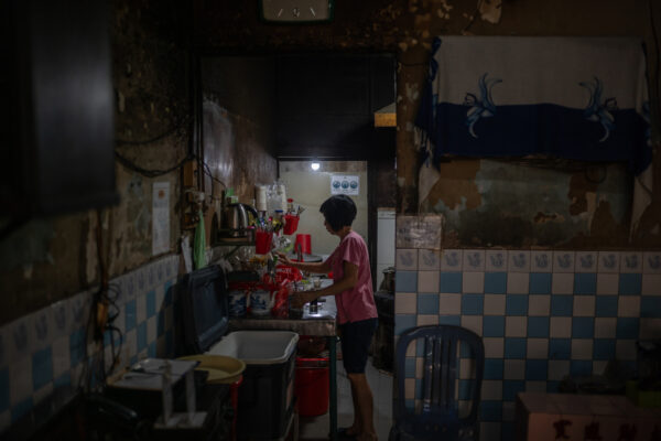 Woman in a pink shirt stands at a cluttered kitchen counter, preparing food in a dim, tiled room.
