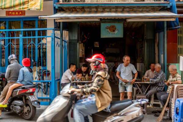 Street-side cafe with older men seated at metal tables while motorcycles pass by on a busy street in front of a blue storefront gate.