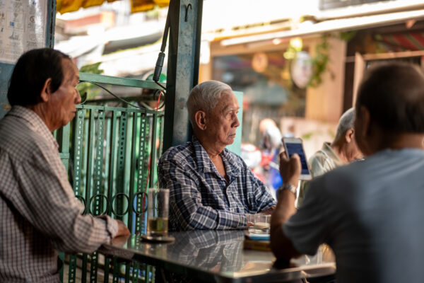 Outdoor cafe scene with three elderly men seated at a metal table; the central man in a plaid shirt looks to the side as another man in the foreground holds up a smartphone.